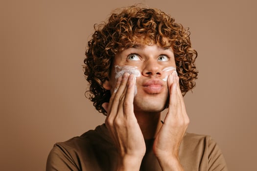 Handsome man enjoying beauty treatment while applying cream on face and standing on brown background photo