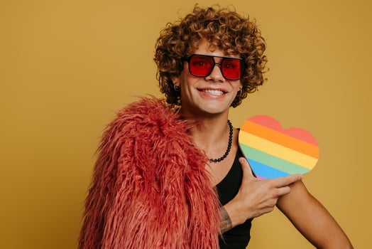 Happy young gay man wearing fluffy coat and holding heart shaped rainbow flag on yellow background photo