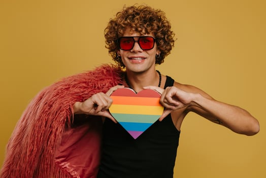 Fashionable young gay man holding heart shaped rainbow flag and smiling on yellow background photo