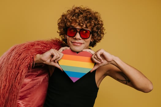 Playful young gay man holding heart shaped rainbow flag and smiling on yellow background photo