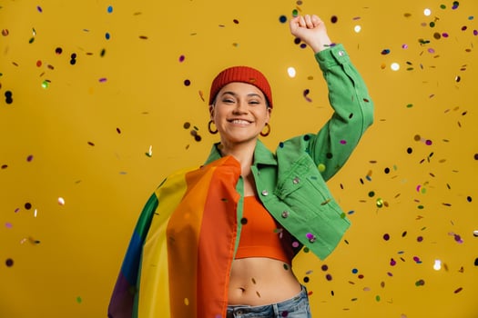Happy woman carrying rainbow flag and gesturing while confetti flying against yellow background photo