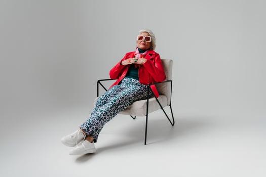 Cool elderly woman in fashionable clothing sitting in comfortable chair on grey background photo