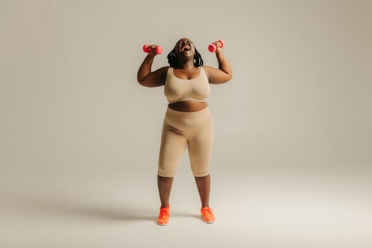 Full length joyful plus size African woman in sportswear exercising with dumbbells on studio background photo