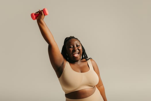 Joyful plus size African woman in sportswear exercising with dumbbell on studio background photo
