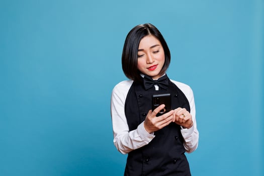 Smiling asian waitress wearing uniform texting on smartphone, managing catering service online. Restaurant woman receptionist typing message on mobile phone, posing in studio photo