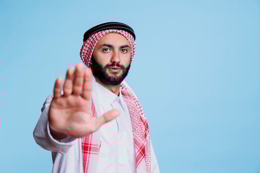 Serious man dressed in muslim thobe and headscarf making stop gesture with arm portrait. Arab person wearing traditional clothes making with prohibition with hand and looking at camera photo