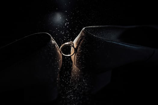 A ring is being shown in a dark room. The ring is gold and is being held by a person. Scene is romantic and intimate photo