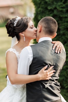 A bride and groom are kissing in a garden. The bride is wearing a white dress and the groom is wearing a black vest photo