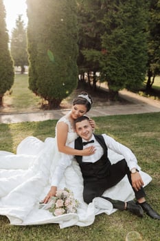 A bride and groom are sitting on a blanket in a park. The bride is wearing a white dress and the groom is wearing a black suit. They are holding hands and seem to be enjoying each other's company photo