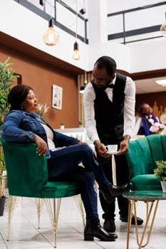 African American woman traveler ordering coffee while resting in comfortable hotel lounge area, sitting in armchair talking with waiter. Providing good customer service in hospitality industry photo