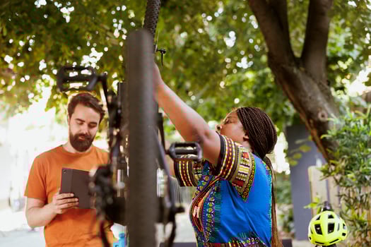 Caucasian man standing with digital device while african american woman repairs damaged bike. Multiethnic couple using phone tablet for researching of bicycle adjustments for summer maintenance at home yard. photo