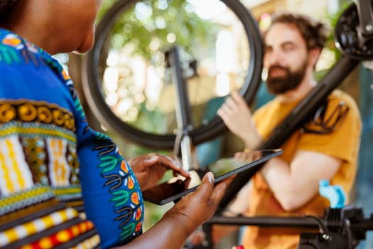 Young african american female holding phone tablet while man in background inspects bicycle wheel outside. Black woman using smart digital device to look for solutions of damaged bike tire. photo