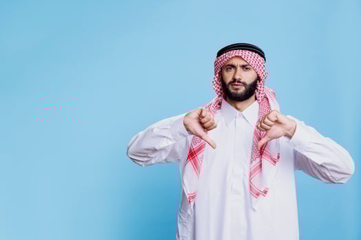 Unhappy muslim man in traditional attire showing two thumbs down disagreement gesture portrait. Discontent arab person showcasing disagreement sign with fingers and looking at camera photo