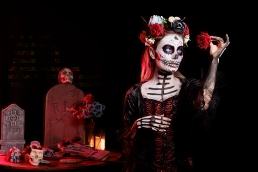 Woman looking at red rose in graveyard, celebrating Day of the Dead. Mysterious lady acting as death goddess, surrounded by frightening funeral items during Halloween horror event photo