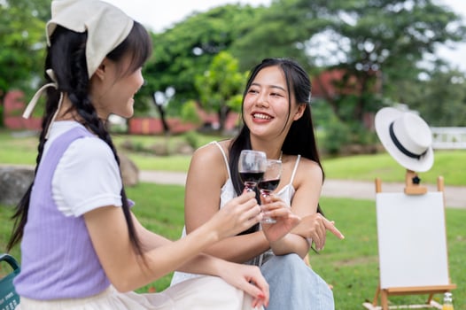 Two happy Asian best friends are clinking glasses of wine while enjoying a picnic in a green park. photo