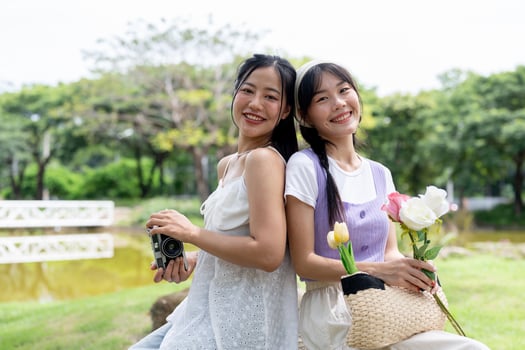 Two lovely Asian woman are sitting on a stone together in a park and smiling at the camera. photo