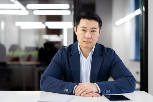 Portrait of a confident Asian businessman seated at a table in a well-lit, contemporary office environment, looking at the camera with focus. photo