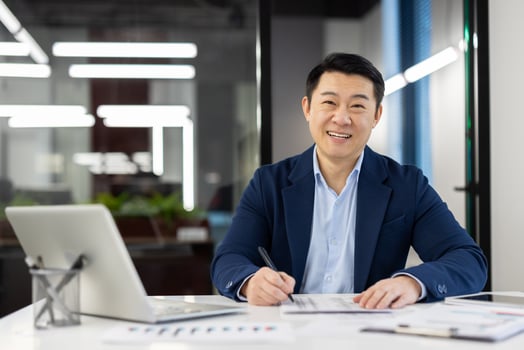 Portrait of a smiling young Asian man in a suit sitting in the office at a desk with a laptop and looking at the camera, working with documents and data. photo