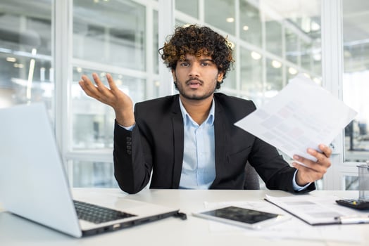 A confused young businessman gesturing with his hand while holding documents, sitting at his office desk, surrounded by laptop and smartphone. photo