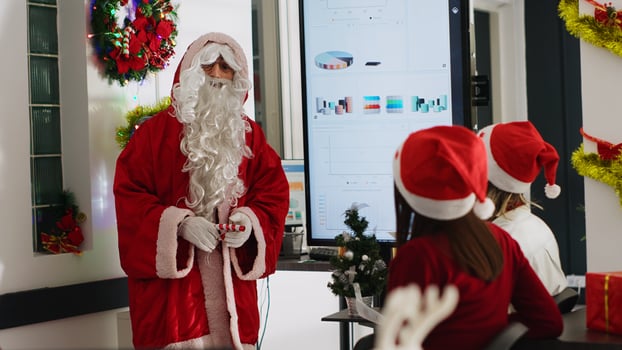 Supervisor dressed as Santa during Christmas season holding team meeting, showing company data on digital screen. Team leader in xmas decorated workplace talking with employees photo