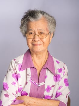 Beautiful elderly Asian woman with short gray hair wearing glasses and looking at the camera with a smile while standing on a gray background. Aged people and health care concept photo