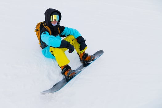 Young man in sportswear and with snowboard sitting on snowy terrain looking towards mountains on cloudy winter day photo