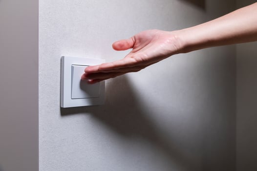 Close-up of an unrecognizable young Caucasian woman's hand turning on a light in a dark room by pressing a switch on a white wall. Close-up photo