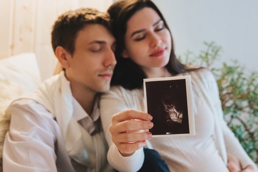 Young happy pregnant woman holding pregnancy ultrasound, showing sonogram picture to pleasantly surprised husband crying from happiness. Family couple waiting for baby photo