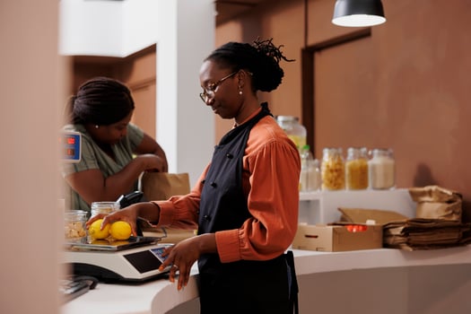 An aproned African American woman assists a customer at a fresh food market with measuring the weight of lemons on digital scale. Female seller weighing locally grown bio produce. photo