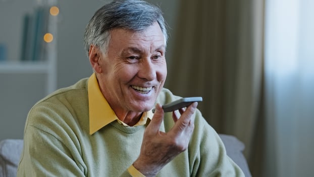 Mature grey-haired 60s man 70s senior old male at home hold smartphone speak loud to phone microphone speaker audio voice message using speakerphone talking with children remote mobile conversation photo