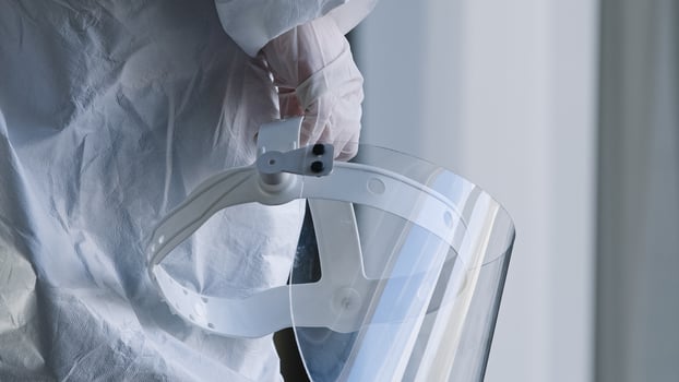 Unrecognizable doctor medical worker nurse practitioner scientist stands in clinic laboratory hospital at window wears white uniform and latex gloves holds in hand transparent protective face shield photo
