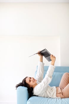 Happy young woman lying down on cozy sofa at home holding open laptop above her with hands up photo