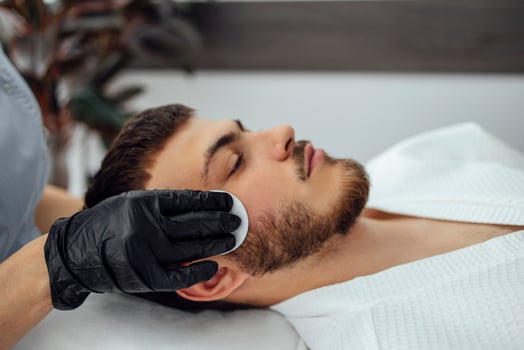 Facial skin cleansing in beauty salon. Beautician in black gloves wipes the face of young man with sponge. photo