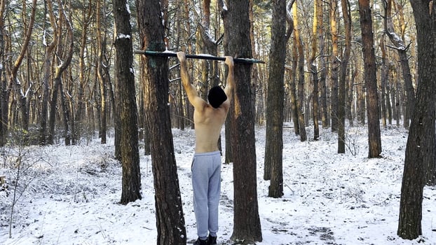 Athletic guy doing pull ups at beautiful forest. Strong and muscular man exercising on horizontal bar at winter nature. Young sportsman training outdoor. Concept of sport and active lifestyle. Slow mo photo