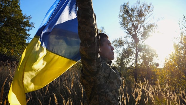 Military man in uniform stands with raised over head flag of Ukraine at countryside. Male ukrainian army soldier with lifted national banner in honor of victory against russian aggression. End of war. photo