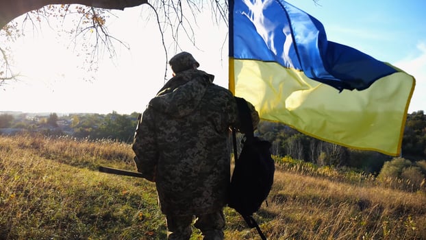 Young male military in uniform walks with waving flag of Ukraine at countryside. Soldier of ukrainian army going with blue-yellow banner outdoor. Concept of victory against russian aggression. photo
