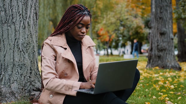 Young focused female student freelancer sitting in autumn park near tree working on laptop typing message checks email studying remote online distance learning using headphones listening to music photo