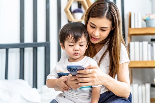 Young asian mother her little adorable son using mobile phone looking at screen enjoying watching funny social media doing online shopping relaxing on bed at home together photo
