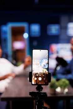 In a neon-lit studio, a vlogger show host is interviewing a guest while a smartphone on a tripod records them. The camera is focused on the phone capturing the content of the influencers. photo