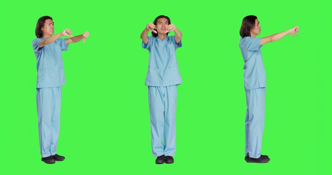 Asian nurse giving thumbs down in studio, showing dislike and negativity symbol over full body greenscreen backdrop. Displeased medical assistant expressing disagreement and disapproval. photo