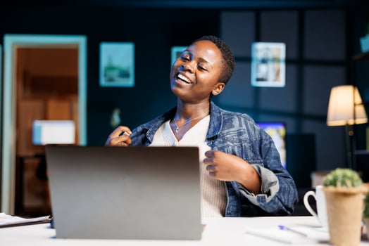 While sitting at her apartment desk, smiling black woman watches comedy film on digital laptop. Enthusiastic African American lady uses personal computer to communicate with friends virtually online. photo