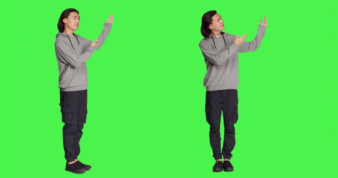 Asian man doing advertisement in studio, pointing at something next to him for new ad slogan. Young person smiling and working on promotional marketing, standing over full body greenscreen backdrop. photo