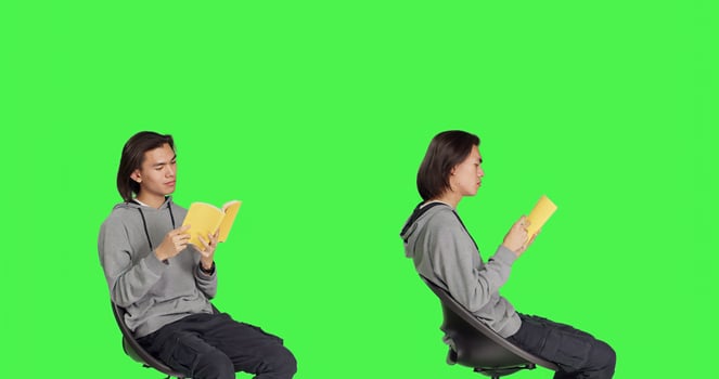 Asian person reading literature book over greenscreen backdrop, enjoying lecture while he sits on chair. Young adult reads fiction novel, being interested in old school hobbies. photo