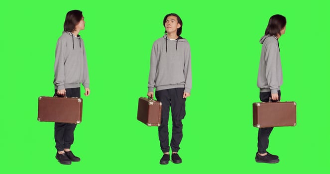 Young adult carries retro suitcase over full body greenscreen backdrop on camera, waiting impatiently for a thing. Asian model holding big suitcase while he checks time on wrist watch. photo