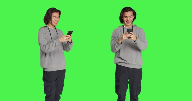 Guy texting messages on mobile phone standing against greenscreen backdrop, using social media app to chat remotely with friends. Asian joyful adult messaging on smartphone. photo