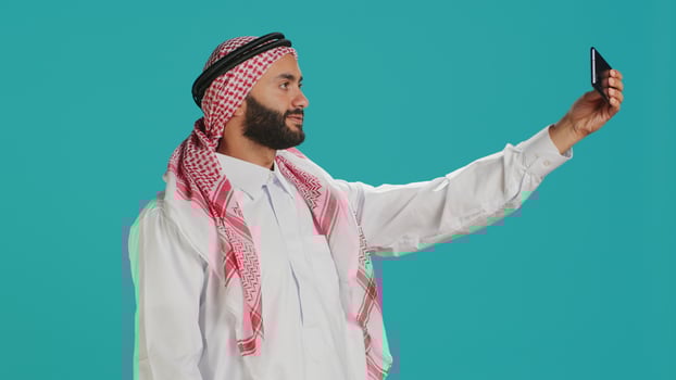 Young arab person takes photos with phone, capturing funny moments on smartphone camera in studio. Middle eastern guy with national headscarf taking pictures in traditional attire.