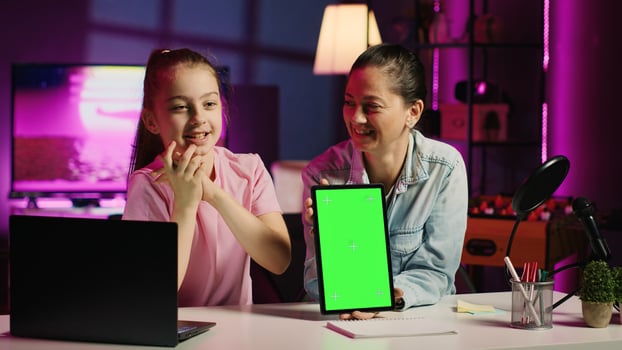 Cute daughter filming internet show helped by her parent, promoting green screen tablet from sponsoring brand. Small child and her mom do influencer marketing, urging fans to purchase mockup device photo