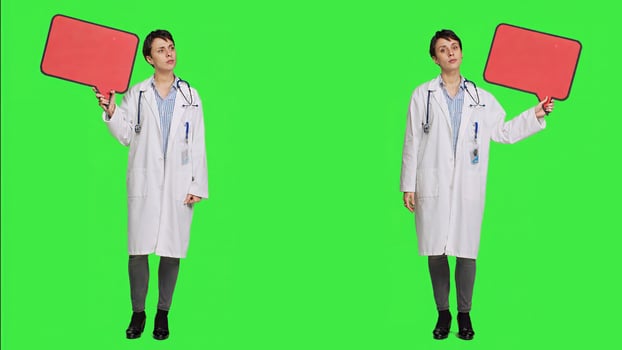 Woman doctor holding a red empty speech bubble sign in studio, showing an isolated blank cardboard icon against greenscreen backdrop. Physician creating announcement with banner. Camera A. photo