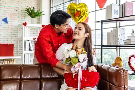 Young asian man or boyfriend surprise and giving flowers at his girlfriend sitting on the sofa at home for the Valentine's day, happy couple with bunch of flowers in house, holidays and people photo
