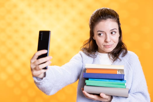 Smiling woman taking phone selfie with stack of books containing information for school exam, studio background. Student taking pictures with smartphone before university assessment, holding textbooks photo